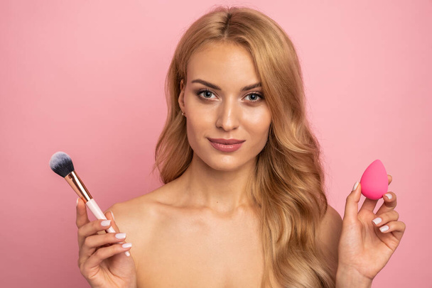 Beauty portrait of a lovely young woman with long blonde hair standing isolated over pink background, using makeup sponge and brush. - Foto, afbeelding