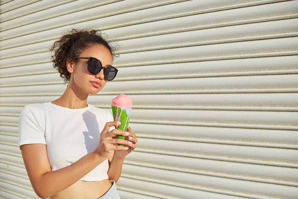 Μια νεαρή όμορφη Αφρο-Αμερικανίδα με λευκό μπλουζάκι και ελαφρύ τζιν τρώει παγωτό στον τοίχο μια ηλιόλουστη μέρα.. - Φωτογραφία, εικόνα