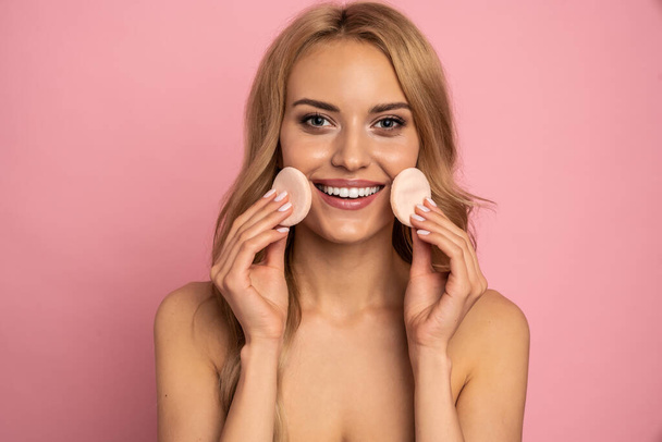 Close up beauty portrait of an attractive smiling young blonde topless woman with bright makeup standing isolated over pink background, holding nakeup sponge. - Fotó, kép