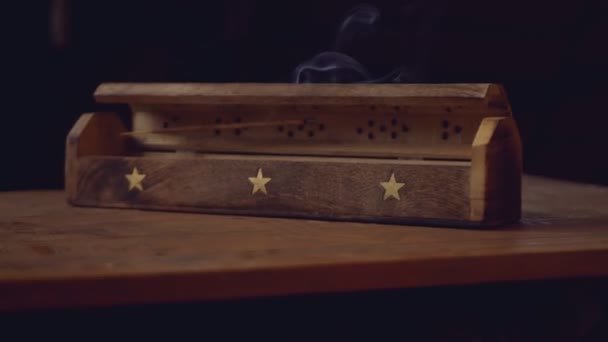 El incienso (varillas de incienso) fuma de una caja de madera única con una estrella. El humo sale de los agujeros. La caja está en una mesa hecha a mano. Rotación en sentido horario. Fondo negro.La mesa levita en el aire
. - Imágenes, Vídeo
