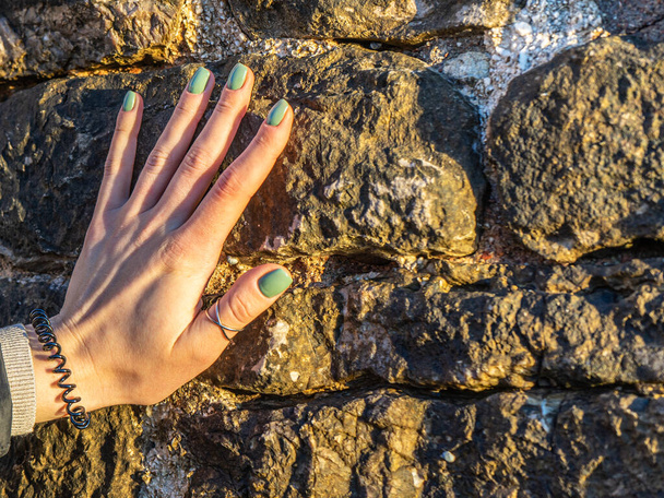 La mano de una mujer con una hermosa manicura verde toca una pared de piedra - Foto, Imagen