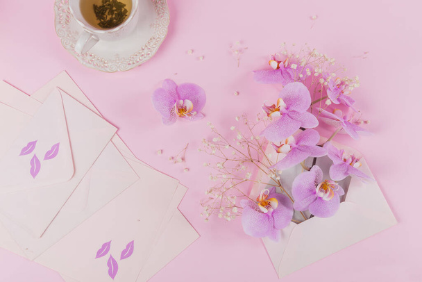 Delicate flatlay composition with morning cup of tea, pink letter bag full of purple orchid flowers and empty envelop on light pink background. Beautiful breakfast surprise concept - Foto, afbeelding