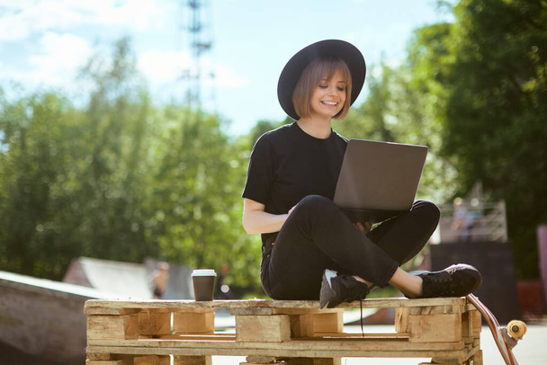 Creative young smiling woman blogger work on laptop on content sitting on wooden pallet with cup of coffee in skatepark - Foto, Imagen