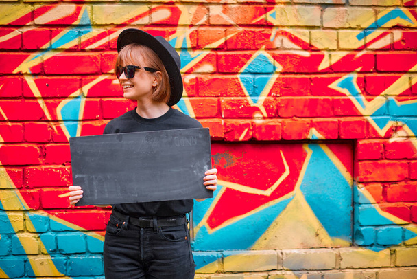 Pretty smiling girl with cardboard sign of protest in hat and sunglasses stand against brick wall with colorful graffiti - Photo, Image