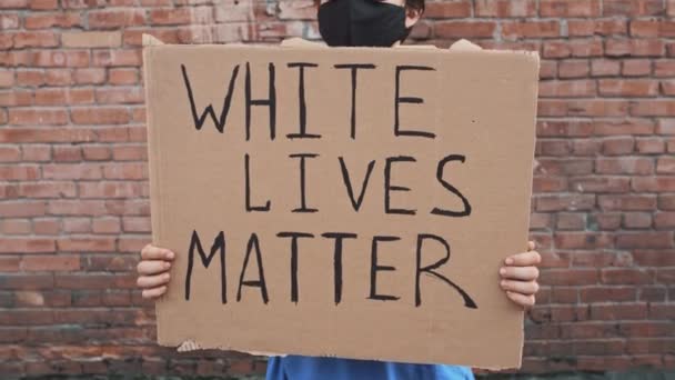 Un homme caucasien masqué se tient contre un mur rouge avec une affiche en carton dans les mains avec une inscription - WHITE LIVES MATTER. Manifestation unique. - Séquence, vidéo