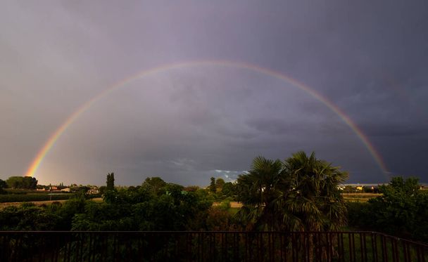 Rainbow with dark clouds and yellow sunbeams and green trees - Foto, Bild