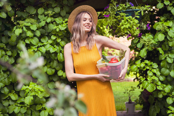 Jeune fille souriante en chapeau dans un jardin vert avec panier de légumes biologiques, photographie pour blog et publicité - Photo, image
