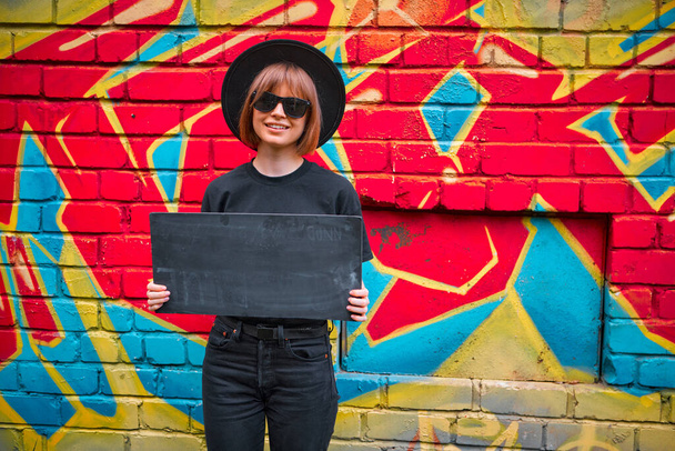 Pretty girl with cardboard sign of protest in hat and sunglasses stand against brick wall with colorful graffiti - Photo, Image