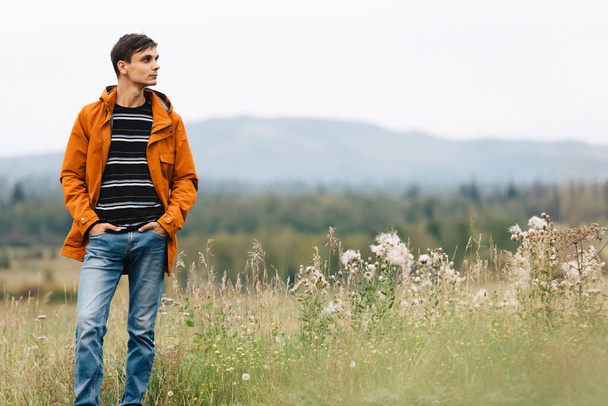 Młody facet w pomarańczowej kurtce, w paski sweter i niebieskie dżinsy stojący na łące na tle jesiennego krajobrazu i spoglądający w bok. - Zdjęcie, obraz