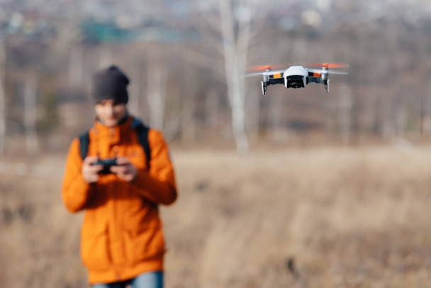 Defocused man in an orange jacket, with a backpack and a hat, controls a quadcopter drone outdoors in autumn. - Photo, Image