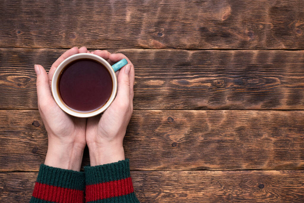 Female hands in sweater with a cup of hot tea above wooden table background with copy space. - Foto, Imagen