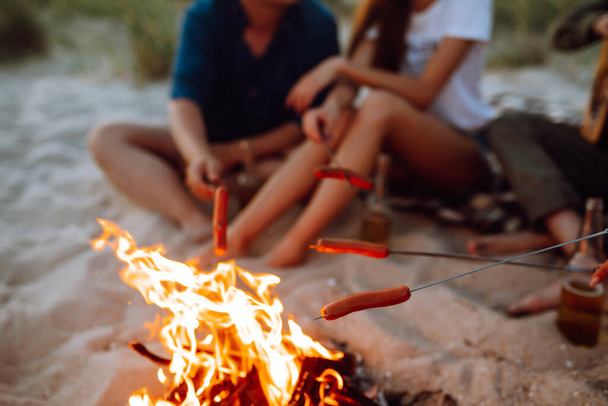 Travel, tourism, hike, picnic and people concept - group of happy friends frying sausages on campfire at the beach. A company of young people came together for a barbecue. - Fotó, kép
