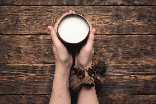 Coconut milk in cup and coconut in female hands on brown wooden table background. - Foto, afbeelding