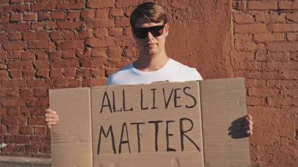 Biały mężczyzna w okularach przeciwsłonecznych i masce stoi pod czerwoną ścianą z kartonowym plakatem w ręku z napisem - ALL LIVES MatTER. Protest pojedynczy. - Materiał filmowy, wideo