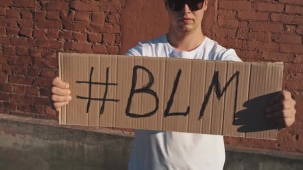 サングラスとマスクの白人男性は、碑文のハッシュタグ- BLMで手に段ボールのポスターと赤い壁に対して立っています。抗議1件. - 映像、動画