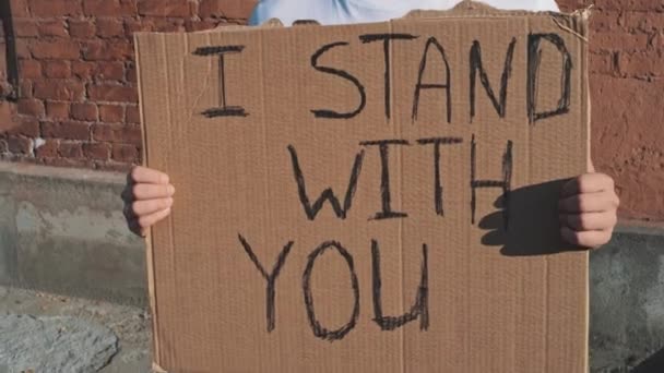Homme caucasien en lunettes de soleil et masque se tient contre le mur rouge avec affiche en carton dans les mains avec inscription - Je STAND WITH YOU. Manifestation unique. - Séquence, vidéo