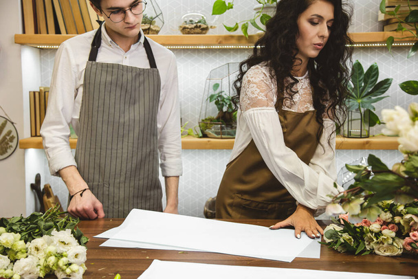 An attractive, experienced female florist teaches a young man how to work in a flower shop. - Photo, Image