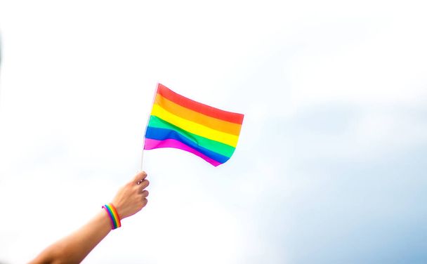  空に虹の旗をつけたゲイのプライドブレスレットを手に。LGBTの人々、同性間の関係、同性愛の概念  - 写真・画像