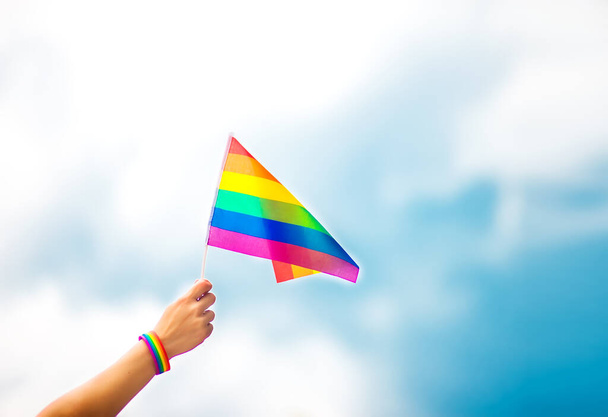  空に虹の旗をつけたゲイのプライドブレスレットを手に。LGBTの人々、同性間の関係、同性愛の概念  - 写真・画像