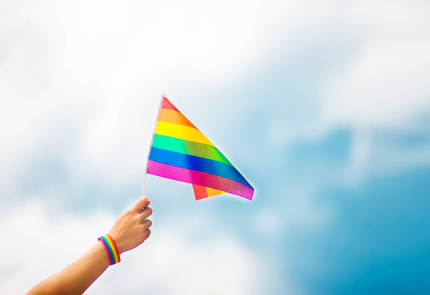  Hand trägt ein Schwulenarmband mit Regenbogenfahnen gegen den Himmel. Homosexuelle Menschen, gleichgeschlechtliche Beziehungen und homosexuelle Konzepte  - Foto, Bild