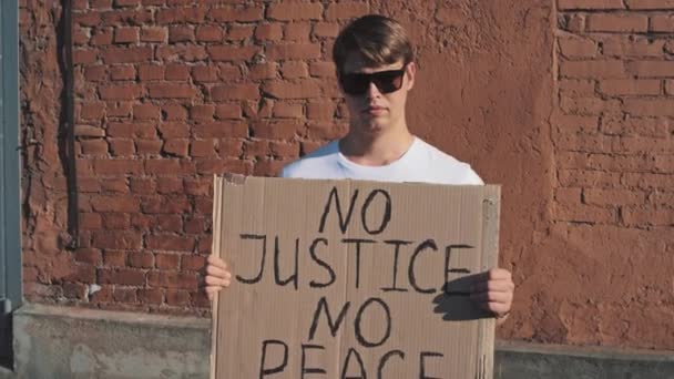 Homme caucasien en lunettes de soleil et masque se tient contre le mur rouge avec affiche en carton dans les mains avec inscription - PAS DE JUSTICE PAS DE PAIX. Manifestation unique. - Séquence, vidéo