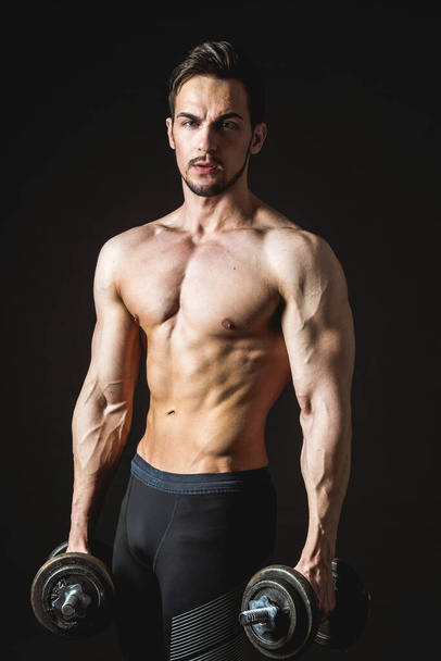 Portrait of an attractive muscular athlete with dumbbells on a dark background - Фото, зображення