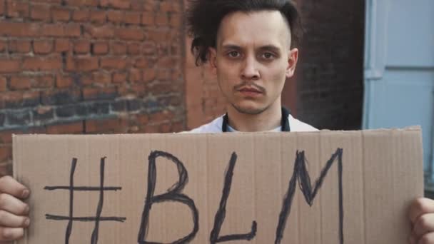 Fehér maszkos férfi áll szemben vörös fal karton poszter a kezében felirat hashtag - BLM. Egyetlen tiltakozás. - Felvétel, videó