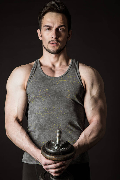 Portrait of an attractive muscular athlete with dumbbells on a dark background - Photo, Image