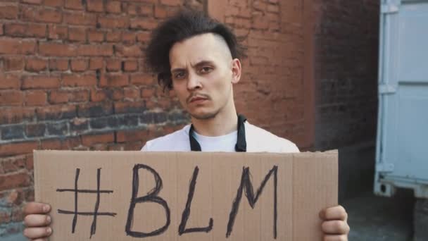 Biały mężczyzna w masce stoi pod czerwoną ścianą z kartonowym plakatem w ręku z hashtagiem z napisem - BLM. Protest pojedynczy. - Materiał filmowy, wideo