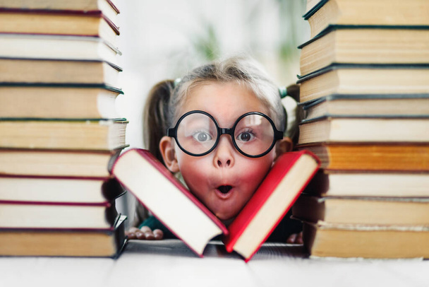 Mädchen im Vorschulalter mit staunendem Gesicht in großer lustiger Brille zwischen einem Stapel Bücher. - Foto, Bild