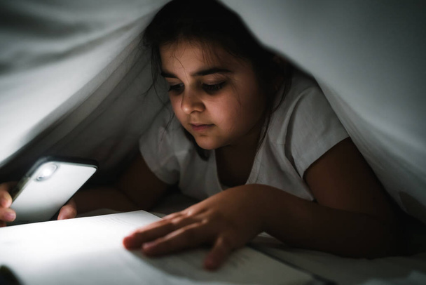 Beautiful Caucasian girl reading under the white sheets with the mobile phone flashlight so she can see - Φωτογραφία, εικόνα