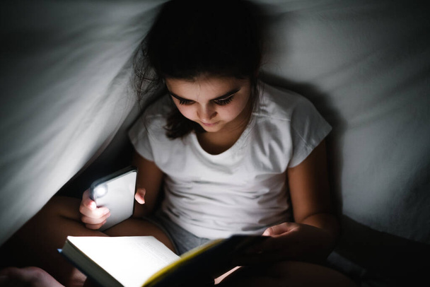 Kaunis valkoihoinen tyttö lukemassa valkoisten lakanoiden alla matkapuhelimen taskulampun kanssa, jotta hän voi nähdä - Valokuva, kuva