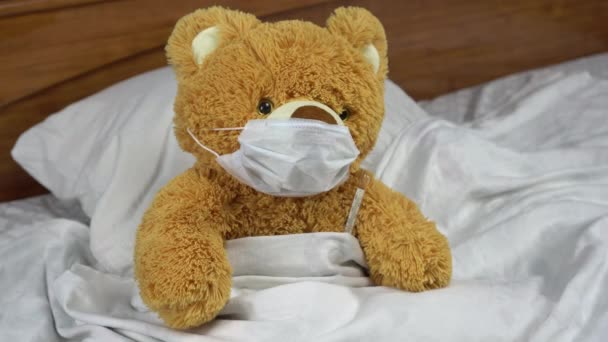 Ein Teddybär misst die Temperatur mit einem Quecksilberthermometer. Der Arzt nimmt das Thermometer. Der Bär liegt mit einer medizinischen Maske im Bett. - Filmmaterial, Video