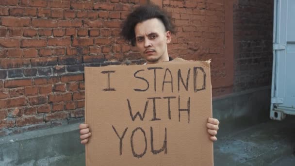 Maskeli beyaz bir adam elinde karton bir posterle kırmızı duvara karşı duruyor. Seninle birlikteyim. Tek protesto. - Video, Çekim