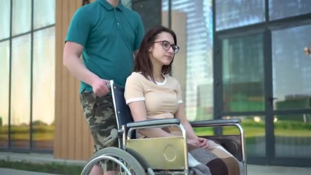 Een jonge man draagt een jonge vrouw in een rolstoel. Een assistent draagt een gehandicapte in een rolstoel langs de straat. Speciaal vervoer voor gehandicapten. - Video