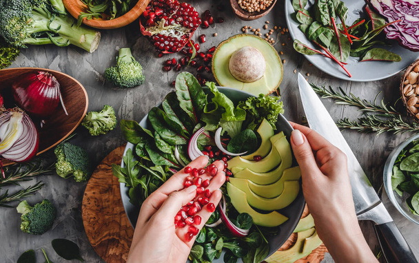 Kadın avokado, yeşillik, roka ve gri arka planda ıspanak ile sağlıklı taze salata pişiriyor. Sağlıklı vejetaryen yemekleri, temiz yemek, perhiz, üst manzara. - Fotoğraf, Görsel