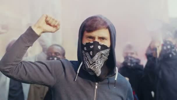 Close up de caucasiano bonito jovem de capuz e cachecol no rosto protestando e gritando slogans em fumaça e gás fora. Rapazes multiétnicos atacando na rua nos EUA
. - Filmagem, Vídeo