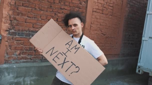 Kaukaski mężczyzna w masce stoi przy czerwonej ścianie z kartonowym plakatem w ręku z napisem - AM I NEXT. Protest pojedynczy. - Materiał filmowy, wideo