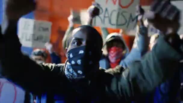 Retrato de un joven manifestante afroamericano con cara enmascarada gritando lemas y gritando consignas. Un tipo protestando por la noche en multitud contra el racismo. Hombre rebelde en motín contra agresión policial
. - Metraje, vídeo