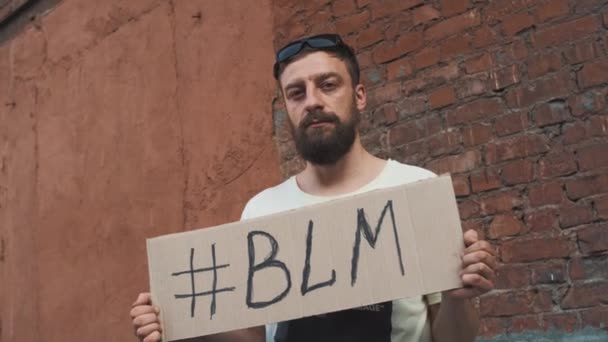 Szakállas fehér maszkos férfi áll szemben vörös fal karton poszter a kezében felirat hashtag - BLM. Egyetlen tiltakozás. - Felvétel, videó