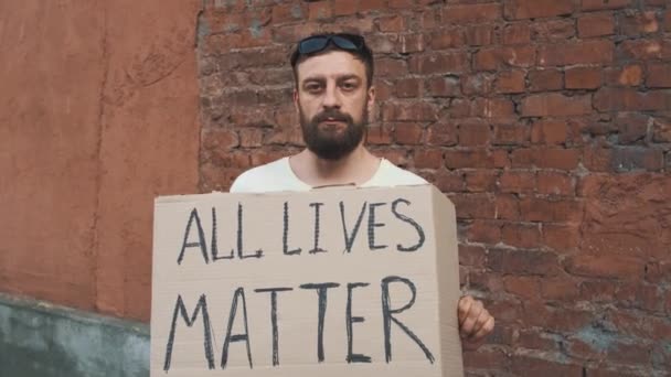Bearded caucasian man in mask stands against red wall with cardboard poster in hands with inscription - ALL LIVES MATTER. Single protest. - Footage, Video