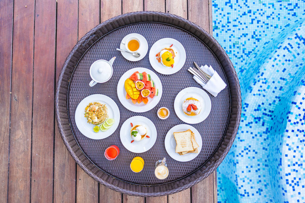 屋外スイミングプールの周りに浮かぶ朝食トレイパンフルーツ卵コーヒーカップとジュース - 写真・画像