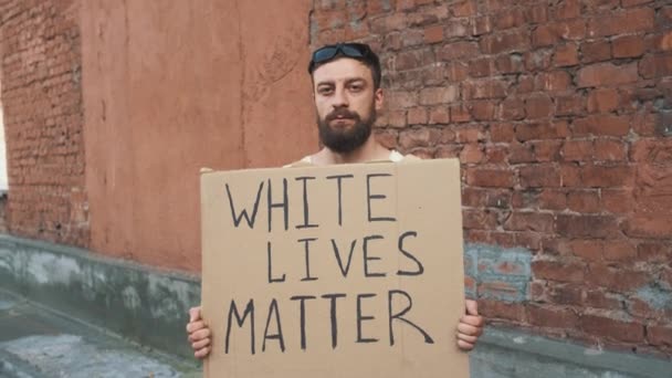 仮面の髭を生やした白人男性は赤い壁に立ち、手にはボール紙のポスターが刻印されています。抗議1件. - 映像、動画