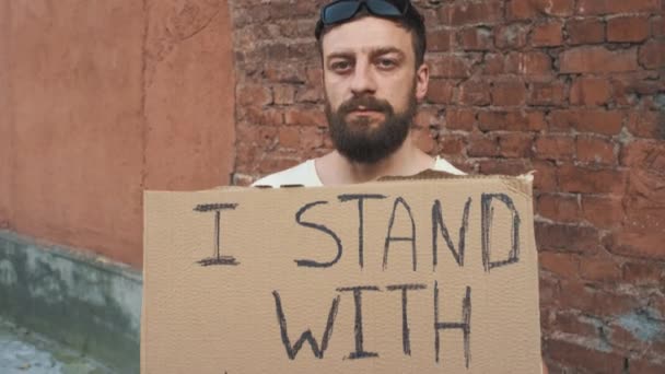 Un homme caucasien masqué se tient contre un mur rouge avec une affiche en carton dans les mains avec une inscription - Je STAND WITH YOU. Manifestation unique. - Séquence, vidéo