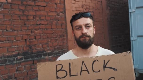 Kaukasischer Mann in Maske steht vor roter Wand mit Pappposter in den Händen mit der Aufschrift - BLACK LIVES MATTER. Einzelner Protest. - Filmmaterial, Video