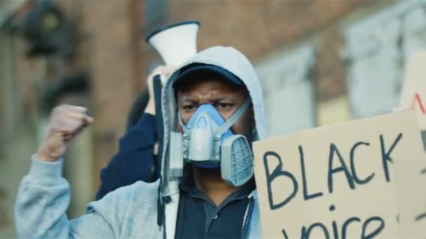 Wieloetniczni młodzi protestujący mężczyźni w maskach i oddychaczach protestujący przeciwko rasizmowi i nielegalnym działaniom policji. Mieszane rasy rebeliantów z plakatami i amerykańską flagą. Mężczyźni krzyczący slogan "Czarne głosy mają znaczenie" - Materiał filmowy, wideo
