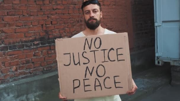 Homme caucasien barbu au masque se dresse contre un mur rouge avec affiche en carton dans les mains avec inscription - PAS DE JUSTICE PAS DE PAIX. Manifestation unique. - Séquence, vidéo