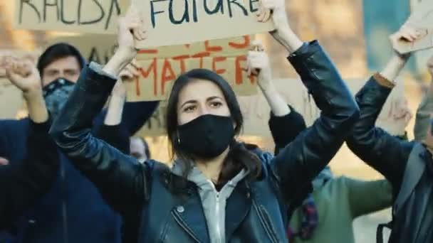 Mooie blanke jonge vrouw in masker hoding poster en schreeuwen motto 's bij protest in multi-etnische mannelijke menigte. Vrouwelijke vrij demonstrant bij manifestatie voor mensenrechten en tegen politiegeweld. - Video