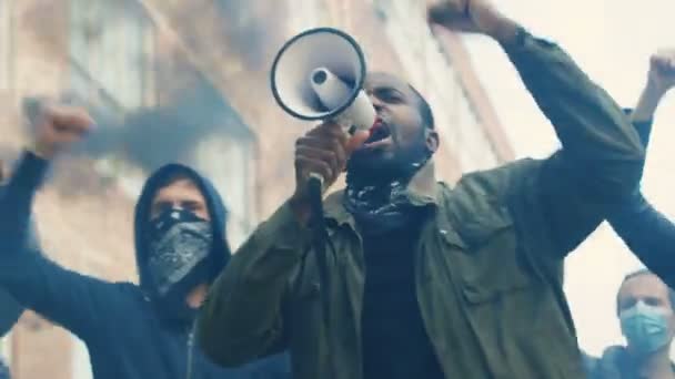 Молодой афроамериканец выкрикивает лозунги в мегафоне на улице в толпе и курит в знак протеста против расизма в США. Агрессивные многонациональные повстанцы выкрикивают свои требования
. - Кадры, видео
