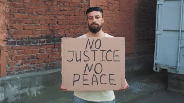 Brodaty mężczyzna kaukaski w masce stoi pod czerwoną ścianą z kartonowym plakatem w ręku z napisem - NO JUSTICE NO PEACE. Protest pojedynczy. - Materiał filmowy, wideo